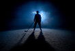 Dark silhouette of a male hockey player in a uniform, helmet and skates with a stick on the ice arena with smoke and blue back light. Sportsman posing at the stadium ice rink. Winter sports.