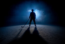 Dark Silhouette Of A Male Hockey Player In A Uniform, Helmet And Skates With A Stick On The Ice Arena With Smoke And Blue Back Light. Sportsman Posing At The Stadium Ice Rink. Winter Sports.