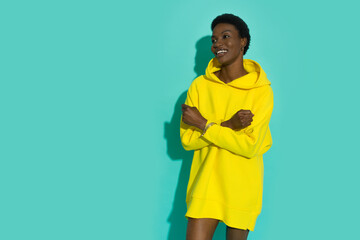 Wall Mural - Smiling young black woman in vibrant yellow oversized hoodie is posing with arms crossed and looking away.