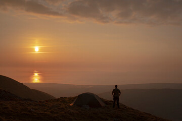 Wall Mural - A man standing beside a wild camping tent at sunset in Snowdonia UK