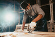 © sutadimages - Confident wood worker expert. Young man working at factory. Skilled carpenter cutting a piece of wood in his woodwork workshop