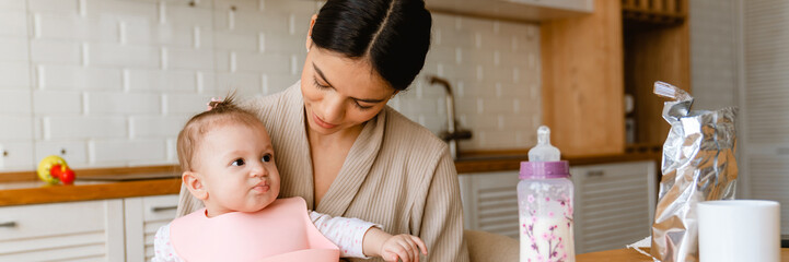 Young brunette mother smiling and feeding her baby in kitchen at home