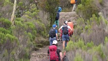 The General Plan Of Porters And Tourists Are Walking Along The Slope Of A Rocky Mountain During A Hike. A Chain Of Porters With Equipment Walking Along A Narrow Path While Climbing Kilimanjaro.