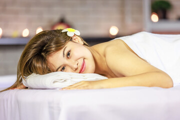 Closeup Asian young beautiful sexy happy female spa customer covered by white clean bath towel laying down resting relaxing closed eyes smiling on massaging bed putting bloom orchid flower on ear