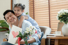 Happy Asian Young Father Getting Bunch Of Flowers And Handmade Card Congratulations On Fathers Day From Little Cute Daughter. Dad Is Piggyback Ride Kid With Love In Living Room At Home
