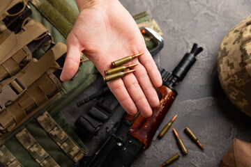 A cartridge in a man's hand against the background of a military body armor, a helmet and a Kalashnikov assault rifle. Composition on a black marble table. Army bulletproof vest. Flat lay