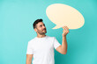 © luismolinero - Young handsome caucasian man isolated on blue background holding an empty speech bubble