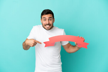 Wall Mural - Young handsome caucasian man isolated on blue background holding an empty placard with surprised expression