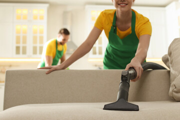 Poster - Professional janitor in uniform vacuuming sofa indoors, closeup. Space for text