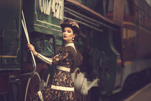 Beautiful Girl In A Historical Retro Dress On A Background Of An Old Steam Locomotive, Steampunk,  At The Railway Station