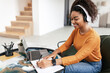 © Prostock-studio - Smiling woman sitting at table, using computer writing in notebook