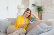 © Prostock-studio - Cheerful young blonde european lady in yellow clothes with wireless headphones looks at phone, sits on sofa