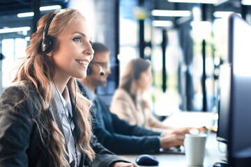 Female customer support operator with headset and smiling, with collegues at background.