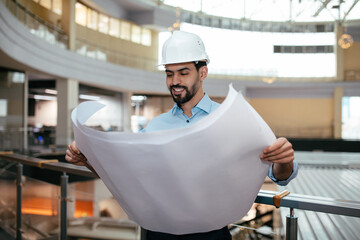 Wall Mural - Happy attractive millennial muslim guy engineer with beard in hardhat checks project drawings in factory