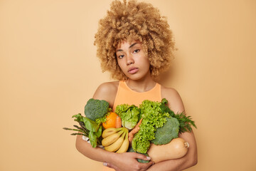 Wall Mural - Studio shot of curly haired young woman embraces healthy fresh fruit and vegetables keeps to diet dressed in casual t shirt isolated over beige background. Green grocery picked from greenhouse