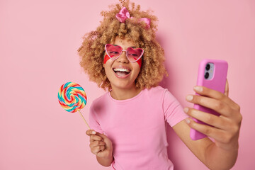 Wall Mural - cheerful curly haired woman holds big rainbow caramel candy on stick laughs gladfully takes selfie v