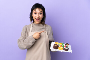 Wall Mural - Young Uruguayan woman holding lots of different mini cakes over isolated purple background pointing finger to the side