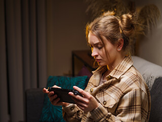 Wall Mural - A pretty blonde girl is playing a video game on a portable game console in the room. Shooting in profile. Room interior. Entertainment, recreation, education.