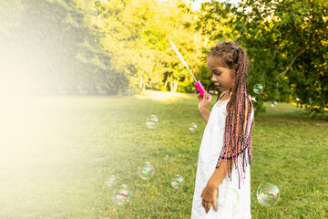 cute girl in white dress and with african pigtails blows soap bubbles in park and have fun. catching