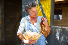 Smiling Mature Farm Owner Carrying Hen And Egg's Bowl In Front Of Chicken Coop