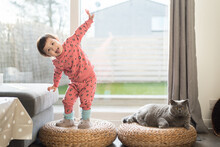 A Toddler Wearing Pink Pyjamas, Long Socks And Slippers Exercises On Top Of A Wicker Stool And Sticks Her Tongue Out While Her British Short Hair Cat Sits Next To Her In A House In Edinburgh, Scotland