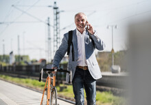 Smiling Businessman Talking On Mobile Phone Walking With Bicycle At Railroad Station Platform