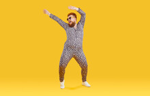 Cheerful Funny Eccentric Fat Man In Good Mood Is Dancing And Fooling Around On Orange Background. Overweight Bearded Man Dressed In Leopard Print Jumpsuit Is Dancing And Fooling Around. Full Length.
