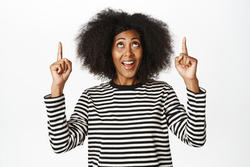 Wall Mural - Enthusiastic female model with afro hair, pointing fingers up and looking amazed at top banner, logo or advertisement on empty space, white background