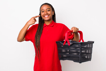 young adult black woman smiling confidently pointing to own broad smile. empty shopping basket concept