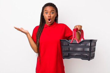 Wall Mural - young adult black woman looking surprised and shocked, with jaw dropped holding an object. empty shopping basket concept