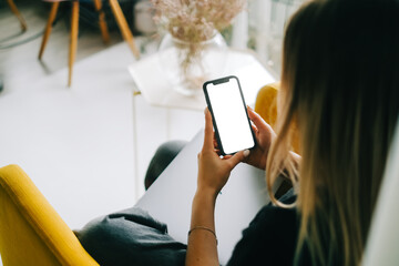 woman holding mobile phone with white screen mock up, resting on a sofa in living room at home.