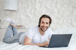 Glad young caucasian man with stubble lies on bed with computer, surfing in internet in light bedroom interior