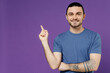 © ViDi Studio - Young smiling fun man 20s wearing basic blue t-shirt point index finger aside on workspace area mock up copy space isolated on plain purple color background studio portrait. People lifestyle concept.
