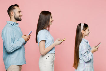 Wall Mural - Side view young happy cheerful fun parents mom dad with child kid daughter teen girl wear blue clothes hold in hand use mobile cell phone isolated on plain pastel pink background. Family day concept