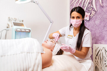 Wall Mural - New normal. Professional female cosmetologist in a protective mask and gloves, applies the mask to the client's face. The concept of professional cosmetology service