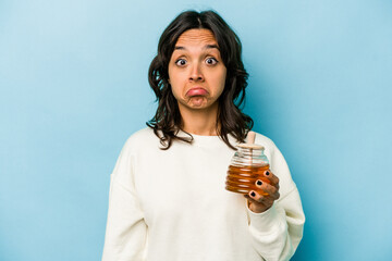 Wall Mural - Young hispanic woman holding a honey isolated on blue background shrugs shoulders and open eyes confused.