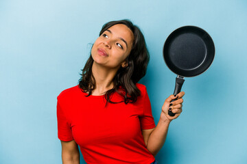 Wall Mural - Young hispanic woman holding a frying pan isolated on blue background dreaming of achieving goals and purposes