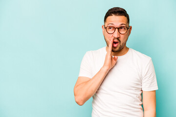 Wall Mural - Young hispanic man isolated on blue background is saying a secret hot braking news and looking aside