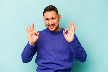 Wall Mural - Young hispanic man isolated on blue background cheerful and confident showing ok gesture.