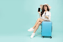 Happy Asian Woman Sitting On Chair With Suitcase And Showing Screen Mobile Phone Isolated On Green Background, Tourist Girl Having Cheerful Holiday Trip Concept
