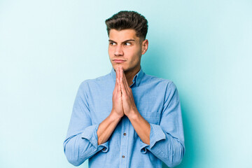 Young caucasian man isolated on blue background praying, showing devotion, religious person looking for divine inspiration.