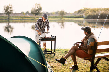 Wall Mural - Senior man preparer food on barbecue grill  while his son is playing acoustic guitar during their camping day in nature.