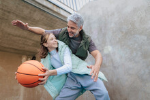 Happy Father And Teenage Daughter Playing Basketball Outside At Court.