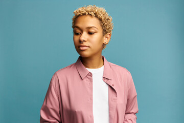 Wall Mural - Beautiful condescending arrogant blonde African woman standing with pensive face expression, raised eyebrows, looking down, dressed in pink shirt isolated against blue background, wearing nose ring