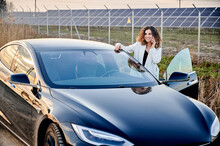 Young Woman Talking On Phone While Standing Near Open Car. On Backgdrop Solar Power Plant Producing Sustainable Electricity And Renewable Energy. Economic And Ecology Energy Indystry.