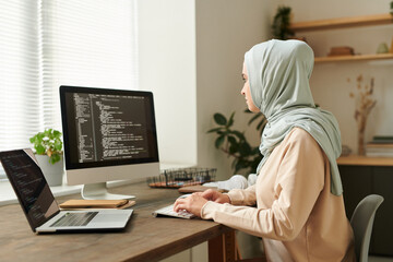 Modern Muslim girl wearing light blue hijab sitting at desk working on new software development, copy space