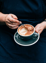Vertical Shot Of A Hand Holding A Saucer And A Cup Of Coffee While Scraping Off The Froth In It.