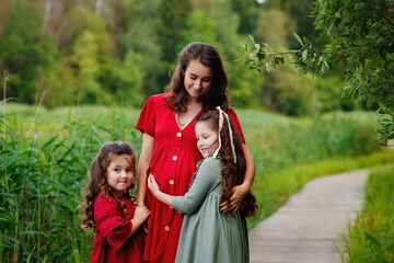 A pregnant mother in a red dress and her two daughters in nature. The family is standing on the bridge holding a bouquet of daisies in their hands.