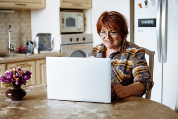 Smiling active senior woman in glasses using laptop, online study in the kitchen. Chatting with family, video calling