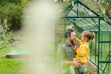 Wall Mural - Father with his little daughter bonding in front of eco greenhouse, sustainable lifestyle.